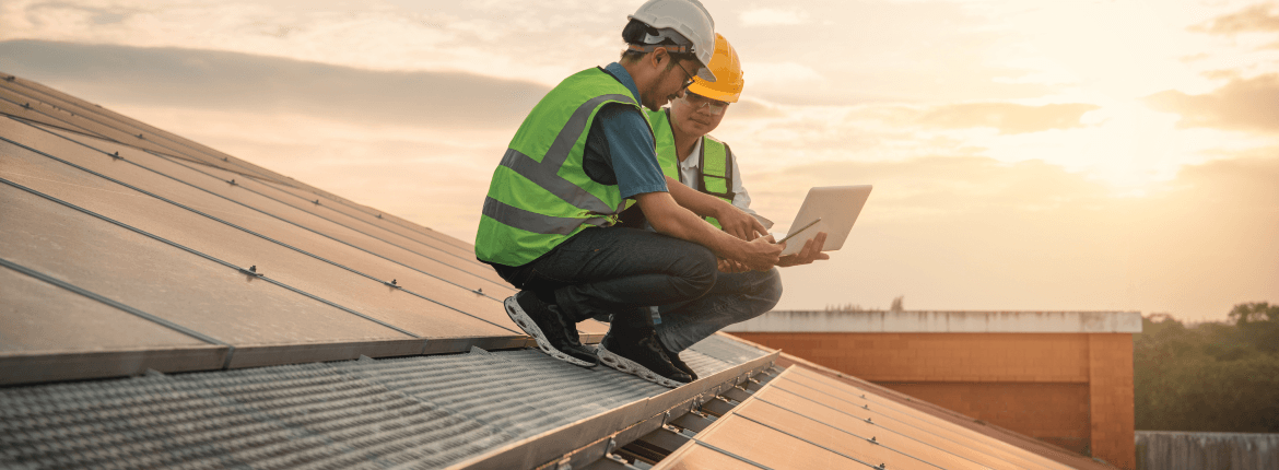 Two roofing and solar system engineers reviewing panel installations using a laptop while also wearing protective gear.