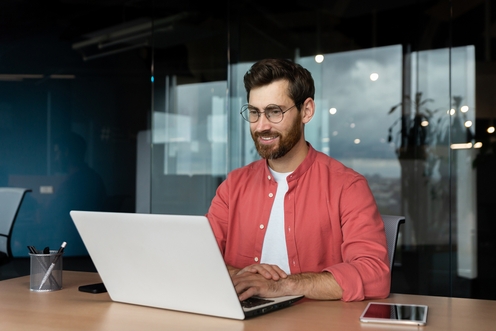 A man with glasses and a beard sits at a desk, focused on his laptop, creating a productive work environment