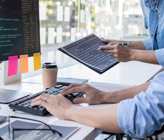 Two individuals collaborating on a computer while reviewing notes on a clipboard