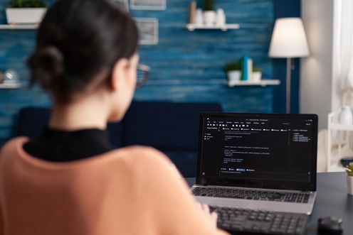 A software developer woman focused on her laptop, with a computer screen displaying data beside her in a modern workspace