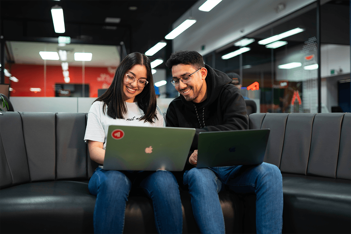 Two business professionals seated on a couch, each using a laptop, engaged in a focused discussion or work session.