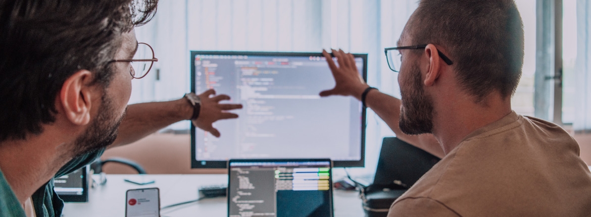 Two male developers reviewing code through a desktop computer monitor and a laptop